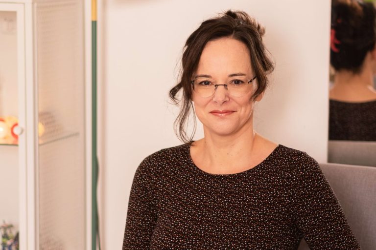 Frau mit Brille und dunkler Bluse lächelt vor hellen, skandinavischen Möbeln.