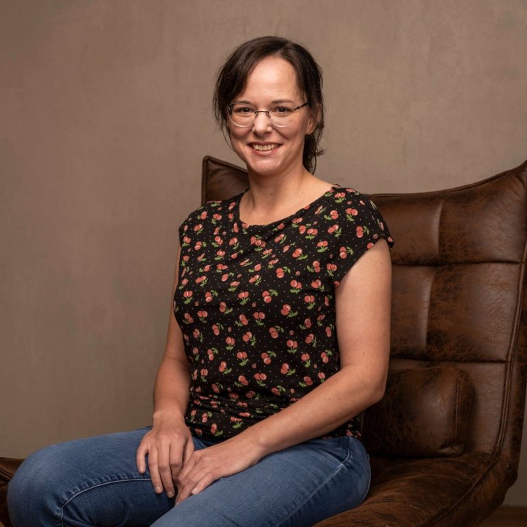 Frau mit Brille sitzt lächelnd auf einem Sessel, trägt ein blütenmusterndes Shirt.