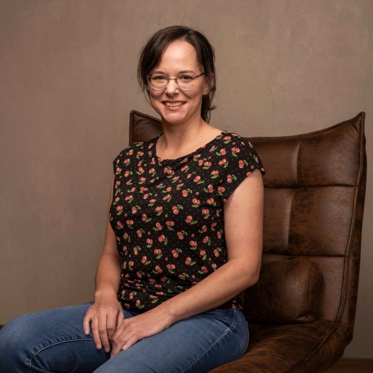 Frau mit Brille sitzt lächelnd auf einem Sessel, trägt ein blütenmusterndes Shirt.