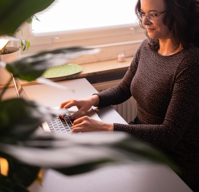 Frau tippt an einem Laptop in einem hellen Raum mit Pflanzen im Hintergrund.